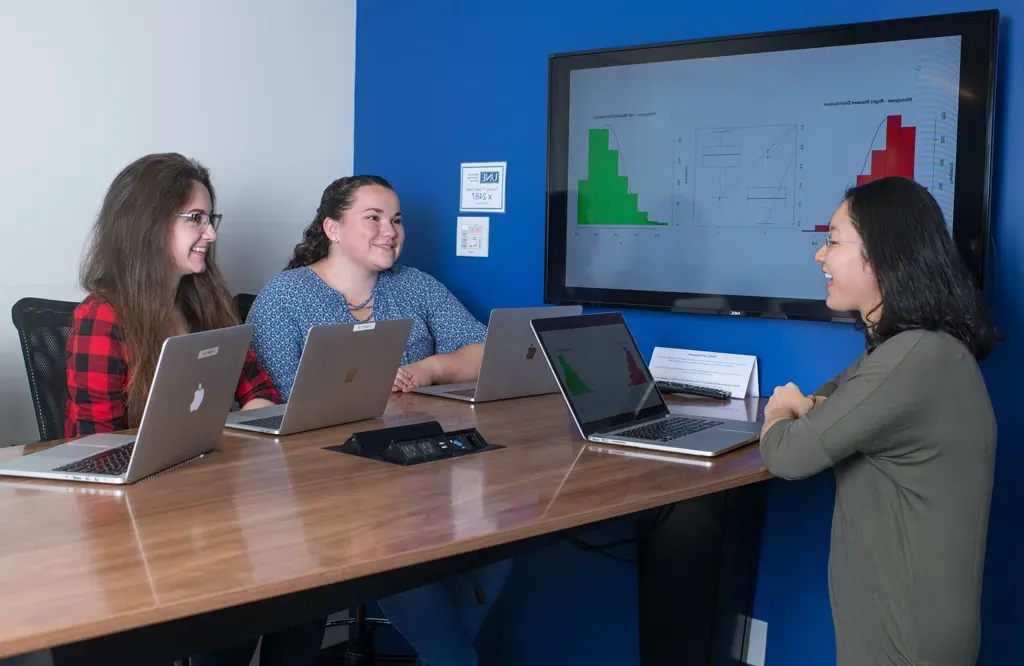 A group of data science students sit at a table with their laptops and graphs on a large monitor